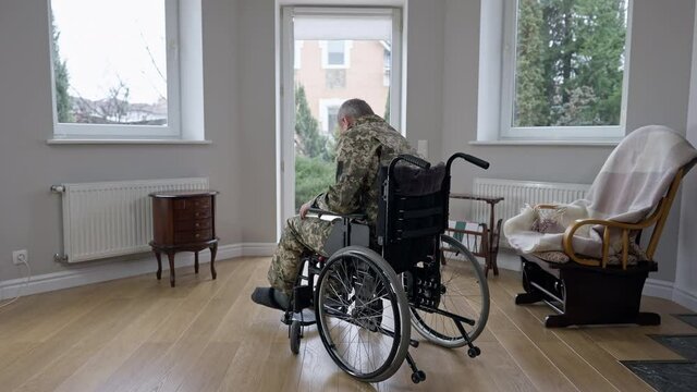 Wide shot back view of motivated grey-haired military veteran trying to stand up from wheelchair. Paraplegic Caucasian man making efforts to recovery at home indoors. Medical paralysis and disability