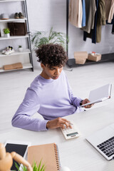 African american retailor using calculator and holding notebook near devices