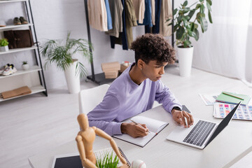 Young african american retailor writing on notebook and using laptop at table