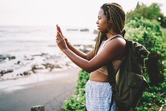 Dark skinned traveler with rucksack making photos of nature environment using smartphone device, millennial tourist with dreads taking pictures via mobile technology connecting to 4g during trip