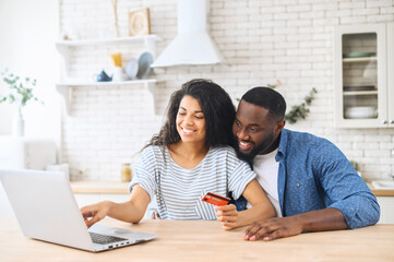 Cheerful African-American couple with laptop shopping online together, beautiful biracial woman holds credit card and points at laptop screen, a guy is agreeing, spouses making online order together