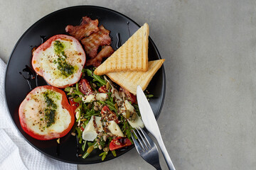 Healthy and tasty breakfast. Fried Eggs in Red Pepper rings, bacon and salad with arugula, cherry tomatoes, seeds and hard cheese on black plate