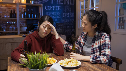 Friendship support lifestyle concept. two young asian chinese women with upset emotion relax sitting in bar at night together. frowning girl comfort sad friend while touching shoulder drinking beer