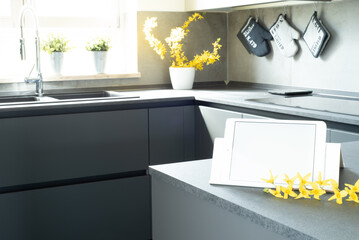 View of modern style kitchen. Mockup of a tablet with white blank screen over the kitchen table with yellow flowers. In the background, window illuminated by natural sunlight. Italian interior design