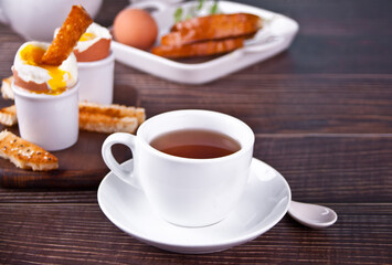 Cup of tea and boiled egg in eggcup on wooden board with crispy toast. Breakfast concept.