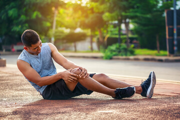 Asian male runner sits on the floor and grips his knee after an acute knee injury from a jogging in a park.