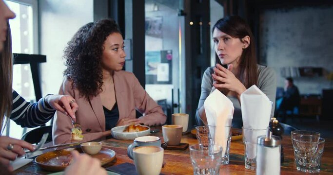 Group of young beautiful happy multiethnic women casually talk at trendy light cafe table with food and coffee at lunch.