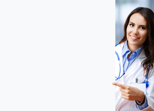 Portrait image of happy smiling female doctor pointing blank signboard with copy space empty area for some text, at office. Young brunette beautiful woman in medical, clinic, healthcare concept