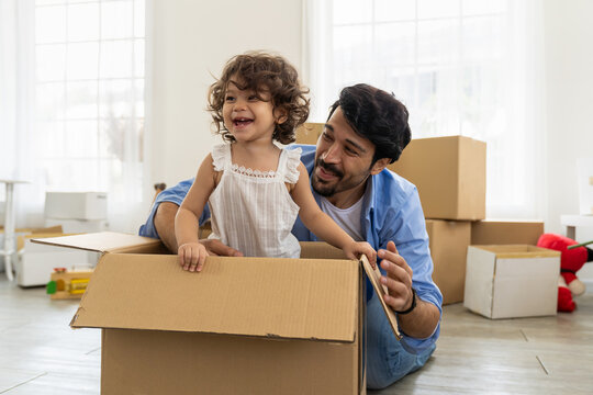 Cheerful father and his little toddler daughter playing inside cardboard box while moving into new home with cardboard boxes on background