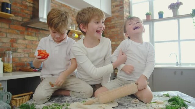 Family with three kids making homemade pizza in a mess, boy eating tomato and its fall on pants, two boys screaming and laughing, boy roll dough in the apartment.