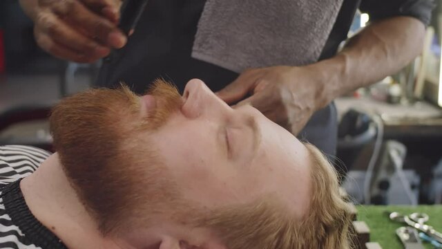Close up shot of young bearded man resting with eyes closed while barber combing and trimming his mustache with scissors in barbershop