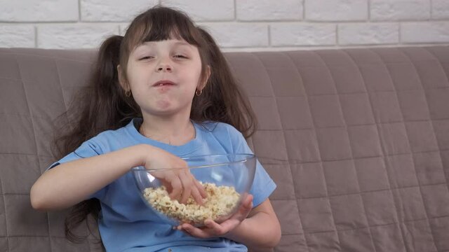 The child is watching TV. A little girl is eating popcorn and watching TV.
