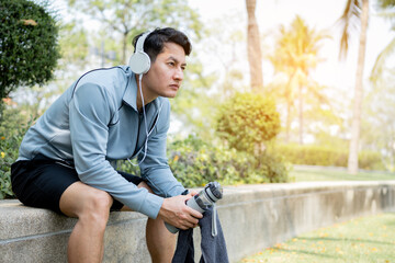 Man runner is sitting and holding bottle of water with headphones. Tired man is drinking water after run exercise.