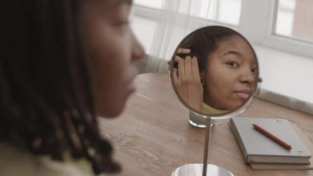Back view of young African-American woman with problem skin and dreads sitting in front of table mirror and enjoying her reflection in it