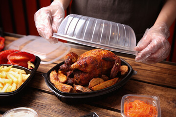 Waiter in gloves closing container with fresh prepared meal at wooden table, closeup. Food delivery...