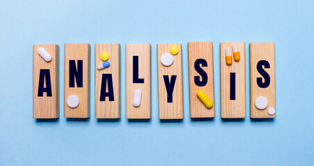 The word ANALYSIS is written on wooden blocks on a light blue table near the pills. Medical concept