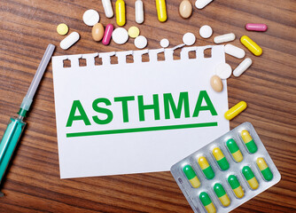 On a wooden table, a syringe, pills and a sheet of paper with the inscription ASTHMA. Medical...