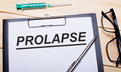 The word PROLAPSE is written on a white piece of paper next to black-rimmed glasses, a pen and a syringe. Medical concept