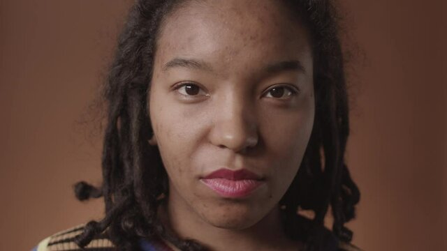 Close up portrait of young African-American woman with problem skin and cool dreads on her head standing against brown background, turning her head and looking at camera