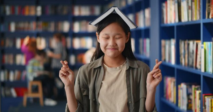 Superstitious asian kid holding school book on head keeping fingers crossed in library