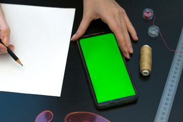 Online sewing training, a woman is taking a course on the phone, female hands, a green screen, a black table, threads and a pattern of a protective mask. Sewing tools