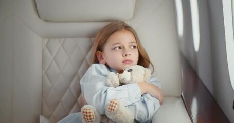 Scared little girl hugging teddy bear sitting in leather chair of business class airplane cabin