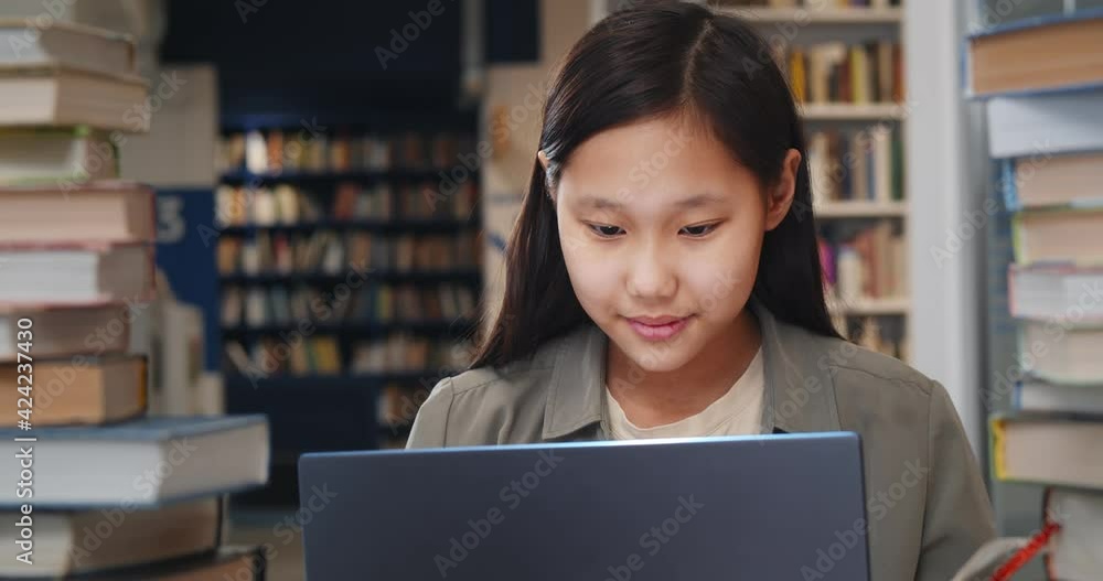 Poster teen asian girl student studying at school library sitting at desk using laptop computer