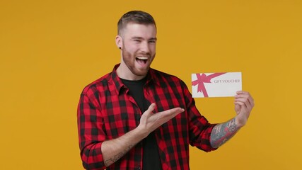 Excited cheerful bearded tattooed young man in red shirt isolated on yellow background studio. People lifestyle concept. Pointing index finger on gift certificate camera on you doing winner gesture