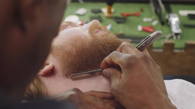 Close up view of Afro-American barber shaving neck of bearded male client with straight edge razor in barbershop