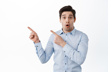 Impressed businessman gasping, say wow and pointing fingers left at copyspace, showing advertisement promo text, standing over white background