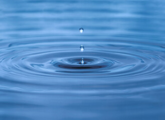 Water droplet dripping into blue pool creating ripples and abstract shape of water. Darkest area in center is in camera focus. Image contains some grain, noise and motion blur