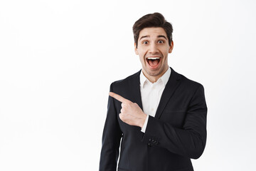 Excited business man, corporate entrepreneur in suit gasping amazed, pointing aside at copyspace with impressed smiling face, checking out awesome deal, white background