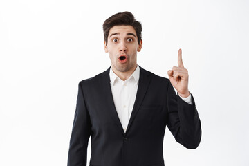 Surprised businessman pointing finger up and gasping amazed, say wow, stare excited at camera, standing in black suit against white studio background
