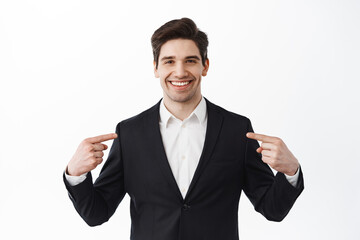 Confident businessman, corporate real estate agent pointing at center, showing logo, self-promoting, standing against white background and smiling