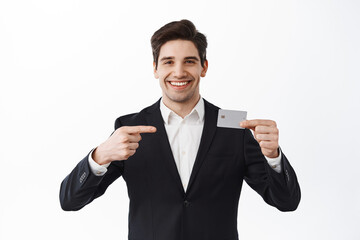Handsome smiling entrepreneur point at credit card, recommend bank, promoting new feature, standing over white background in formal suit