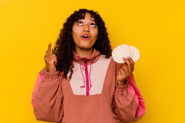 Young mixed race sport woman holding a rice cakes isolated on yellow background pointing upside with opened mouth.