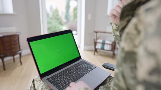 Laptop with green screen on knees of unrecognizable Caucasian military man indoors. Adult soldier or veteran surfing Internet or messaging online at home. Modern technologies, chromakey