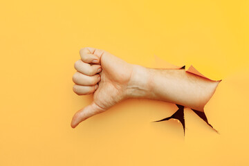 Hand showing a thumb down through ripped hole in yellow paper background. Dislike and disapproval gesture