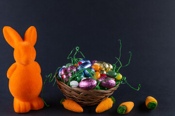 Orangefarbener Hase mit Möhren und einen gefüllten Osternest mit süßen Eiern auf schwarzem Untergrund