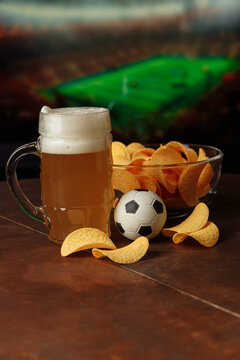 Glass of beer, soccer ball and snack in front of screen with football game. Vertical image