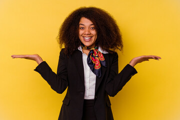 Young African American air hostess isolated on yellow background makes scale with arms, feels happy...