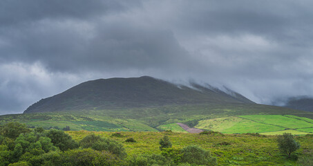 Fototapeta na wymiar Green fields and forest at foothill of Carrauntoohil mountain, hidden in mist or cloud, dramatic storm sky in Blackvalley, Rink of Kerry, Ireland
