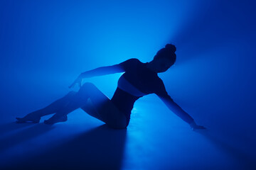 Fototapeta na wymiar Ballerina in black bodysuit dancing with blue light and smoke. Silhouette of dancer in ballet shoes.