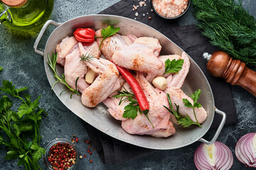 Raw chicken wings in metal pan or bowl with spices and ingredients for cooking on dark grey slate, stone or concrete background. Raw meat with spices at black table. Top view. Mock up.