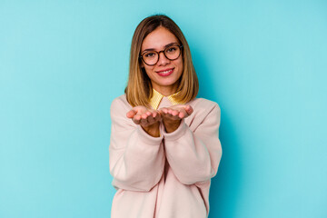 Young student caucasian woman isolated on blue background holding something with palms, offering to camera.