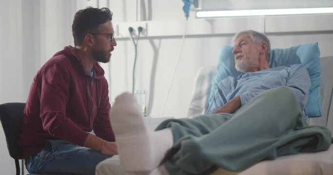 Adult man visiting aged father with broken leg in hospital ward