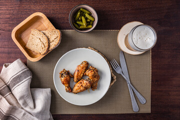 top view of a plate with roasted chicken wings, bowl of pickles, bread slices and pint of beer