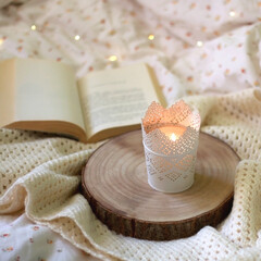 Candle holder with lit candle, open book, soft blanket and bokeh lights on the bed. Cozy details at home. Selective focus.