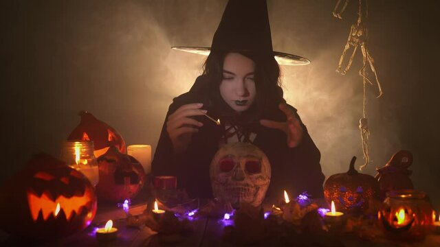 Woman in the image of witch is conjuring over the skull with a joss stick and laughing ominously on black background. A lady in dark mantle surrounded by smoke and Halloween decorations is doing magic