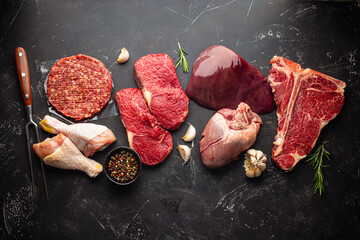 Selection of assorted raw meat food with seasonings for zero carb carnivore diet: uncooked beef steak, ground meat patty, heart, liver and chicken legs on black stone background from above  - obrazy, fototapety, plakaty